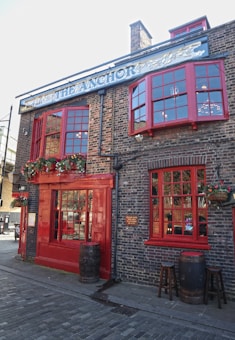 A traditional brick building with large red-framed windows and doors. There are flower pots with greenery and colorful flowers hanging around the entrance and windows. Wooden barrels and stools are placed outside, contributing to a rustic and inviting atmosphere. The sign above the building reads 'The Anchor' with a decorative design.