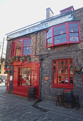 A traditional brick building with large red-framed windows and doors. There are flower pots with greenery and colorful flowers hanging around the entrance and windows. Wooden barrels and stools are placed outside, contributing to a rustic and inviting atmosphere. The sign above the building reads 'The Anchor' with a decorative design.