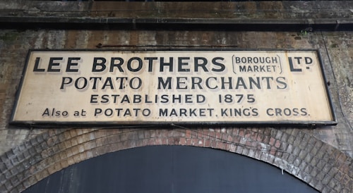 A large, aged sign affixed to an old brick wall reads 'Lee Brothers Potato Merchants Ltd (Borough Market) Established 1875. Also at Potato Market, King's Cross.' The letters are faded and the sign shows signs of wear and discoloration.