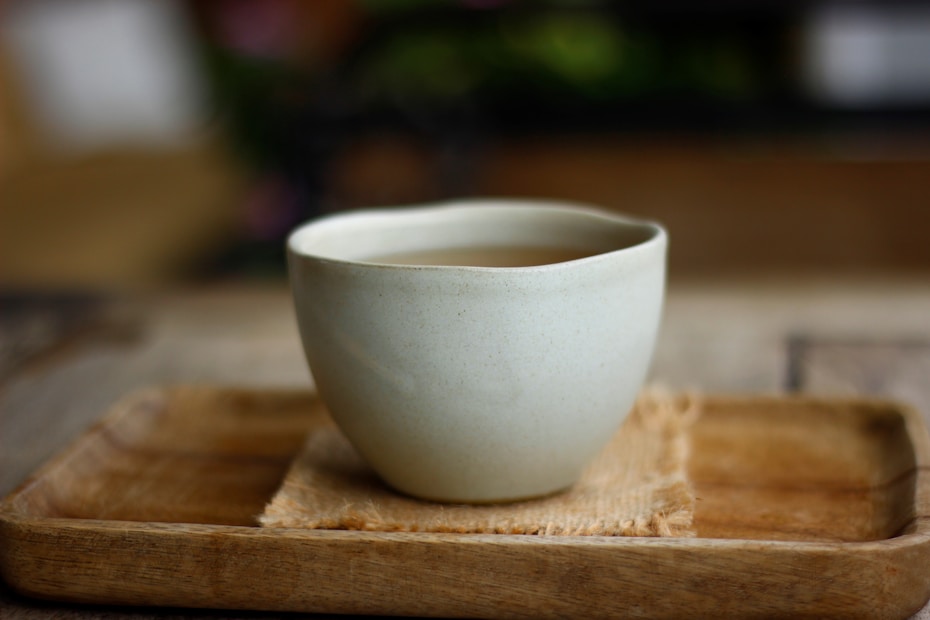 A simple, elegant ceramic cup with a smooth, off-white surface sits on a wooden tray. The tray has a rustic, natural finish and is placed on a woven mat, adding texture. The background is softly blurred, highlighting the cup as the focal point.