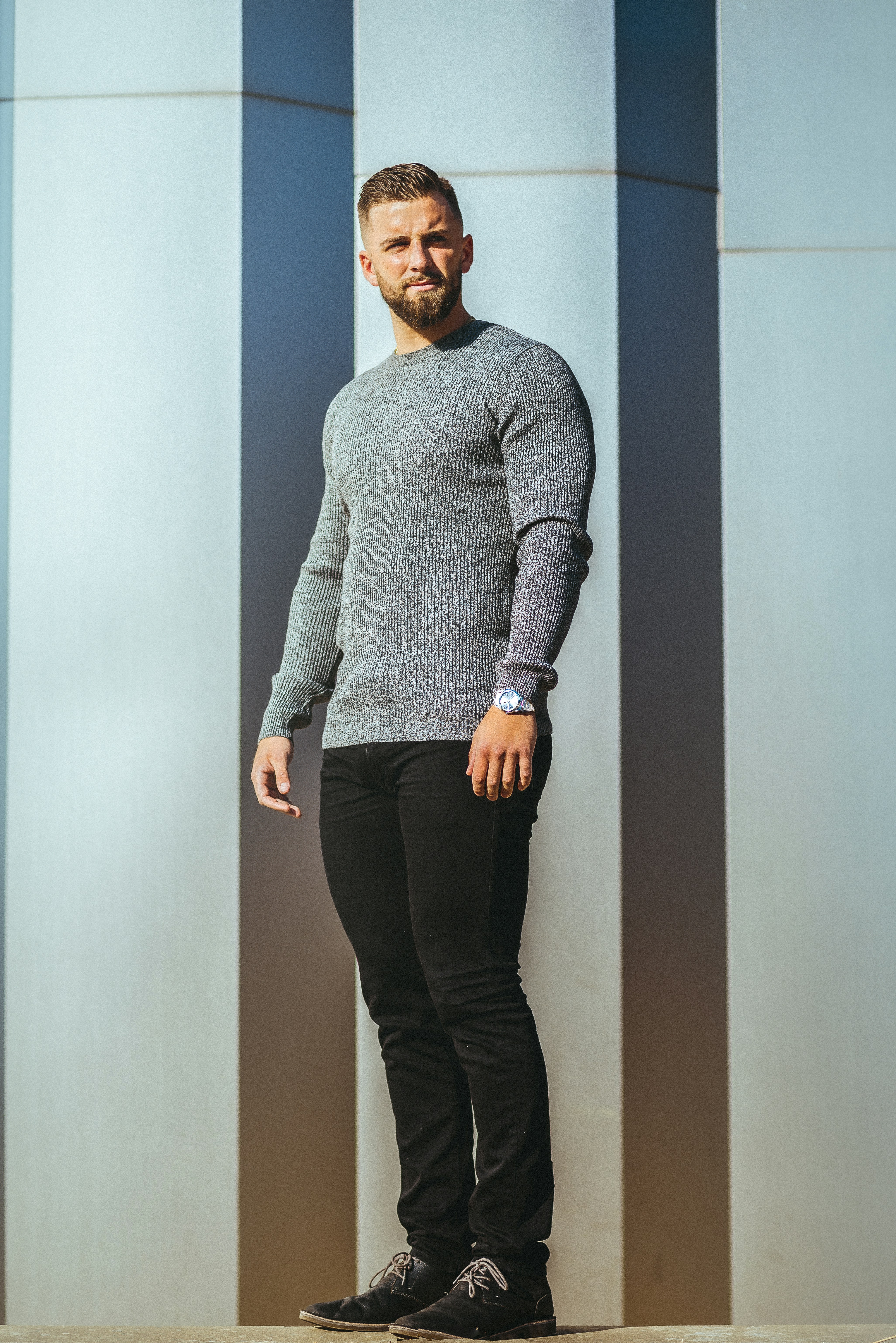 man in gray sweater and black pants standing beside white wall