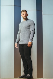 man in gray sweater and black pants standing beside white wall