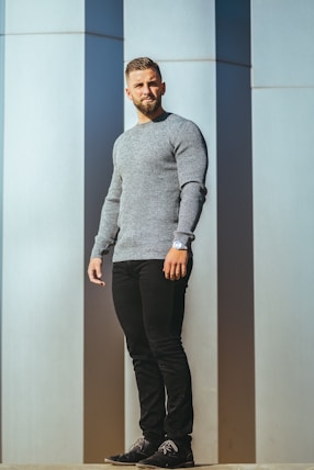 man in gray sweater and black pants standing beside white wall