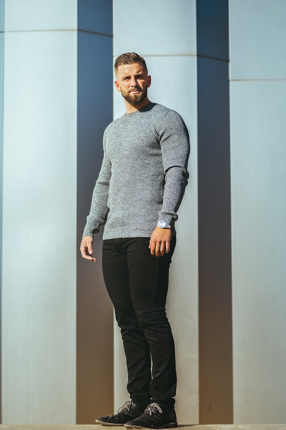 man in gray sweater and black pants standing beside white wall