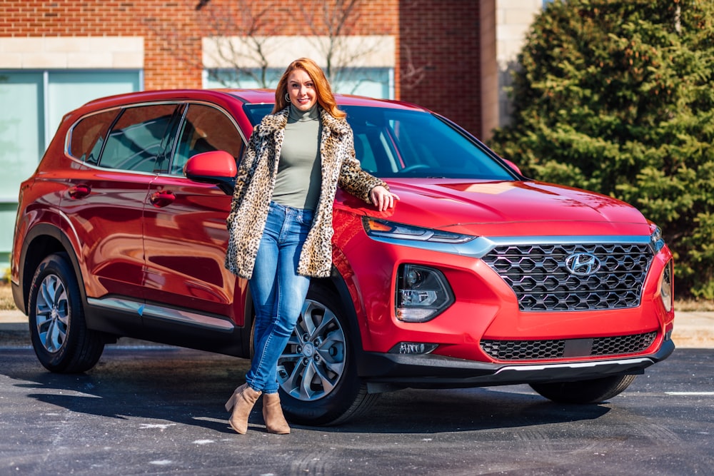 Mujer con bufanda blanca y negra y jeans de mezclilla azules de pie junto a un automóvil Mercedes Benz rojo