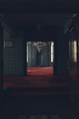 A dimly lit, ornately decorated room with intricately patterned walls and a luxurious red carpet. A large chandelier hangs from above, and windows allow soft natural light to enter. The space appears to be a prayer or meditation area, characterized by its symmetrical and harmonious design.