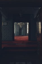 A dimly lit, ornately decorated room with intricately patterned walls and a luxurious red carpet. A large chandelier hangs from above, and windows allow soft natural light to enter. The space appears to be a prayer or meditation area, characterized by its symmetrical and harmonious design.