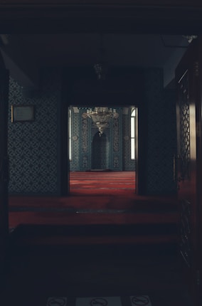 A dimly lit, ornately decorated room with intricately patterned walls and a luxurious red carpet. A large chandelier hangs from above, and windows allow soft natural light to enter. The space appears to be a prayer or meditation area, characterized by its symmetrical and harmonious design.