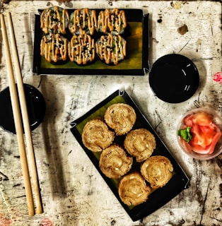 A rustic table setting featuring two trays of sushi rolls. The top tray contains sushi drizzled with a creamy orange sauce and sprinkled with black sesame seeds, while the bottom tray has crispy rolls. Adjacent to the trays are chopsticks and two small black dishes. A small bowl with pickled ginger and wasabi sits nearby.