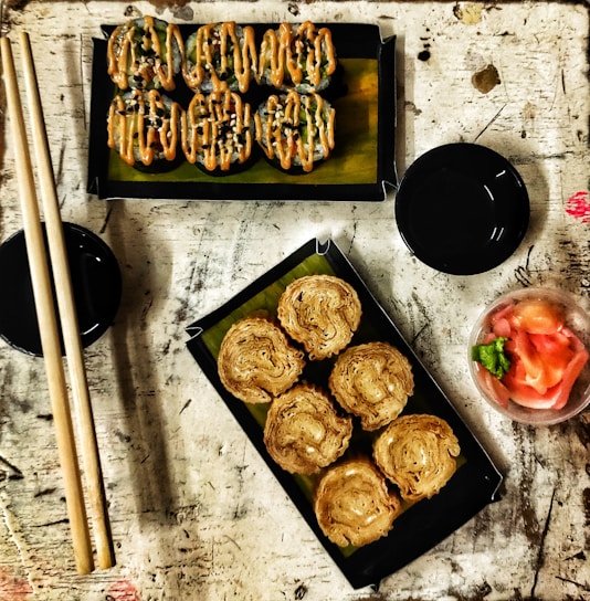 A rustic table setting featuring two trays of sushi rolls. The top tray contains sushi drizzled with a creamy orange sauce and sprinkled with black sesame seeds, while the bottom tray has crispy rolls. Adjacent to the trays are chopsticks and two small black dishes. A small bowl with pickled ginger and wasabi sits nearby.