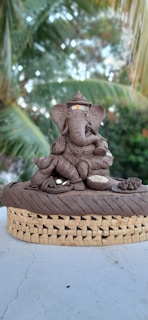 A detailed sculpture of Lord Ganesha made from clay is placed on a woven mat. The sculpture depicts Ganesha with an ornate crown, sitting gracefully with one leg crossed. A small mouse figure, symbolic of his companion, is also present. The background features blurred, lush green palm leaves.