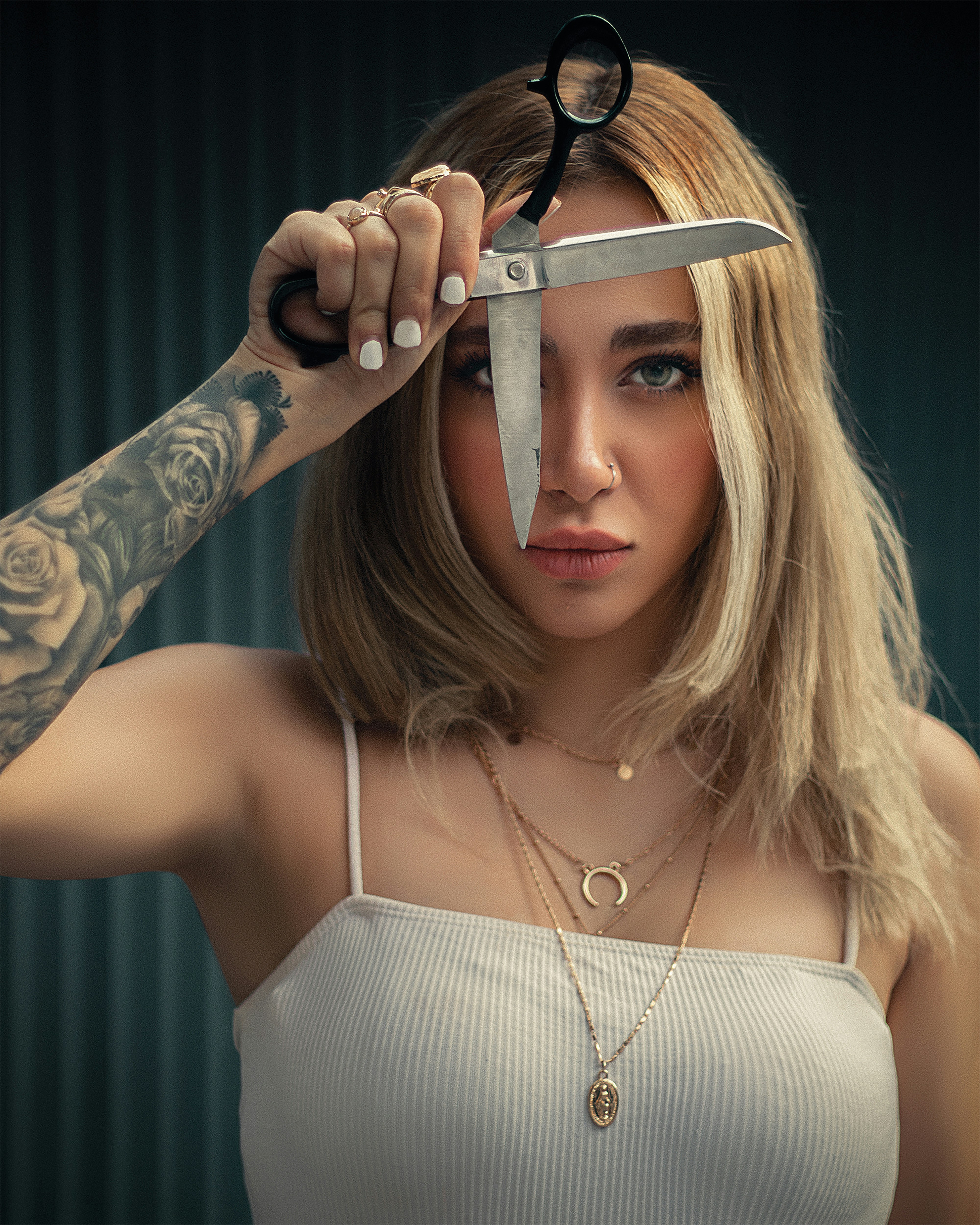 Jess, with blonde hair and tattooed arms, holding open scissors across her face