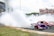 A red and blue sports car is drifting on a racetrack, producing large amounts of white smoke from its tires. A crowd is watching from the stands in the background, and a large screen displays the action. The sky is partly cloudy and there is a patch of grass beside the track.