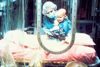 A caregiver wearing surgical attire reads 'The Cat in the Hat' to a child in a protective environment. The child, also wearing protective clothing, smiles while clutching a doll. The setting appears to be a medical isolation room, indicated by the plastic curtains and sterile equipment.