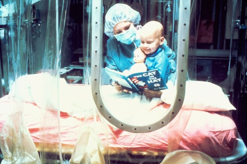 A caregiver wearing surgical attire reads 'The Cat in the Hat' to a child in a protective environment. The child, also wearing protective clothing, smiles while clutching a doll. The setting appears to be a medical isolation room, indicated by the plastic curtains and sterile equipment.