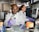 Two scientists in a laboratory, both wearing white lab coats and protective eyewear. The scientist in the foreground is using a pipette, while the one in the background is working on a computer. Various laboratory equipment and supplies are visible around them.