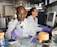 Two scientists in a laboratory, both wearing white lab coats and protective eyewear. The scientist in the foreground is using a pipette, while the one in the background is working on a computer. Various laboratory equipment and supplies are visible around them.