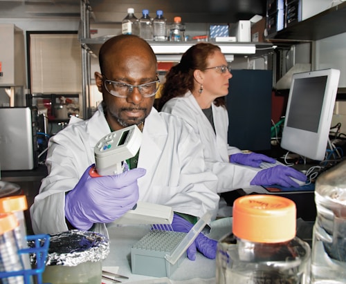 Two scientists in a laboratory, both wearing white lab coats and protective eyewear. The scientist in the foreground is using a pipette, while the one in the background is working on a computer. Various laboratory equipment and supplies are visible around them.