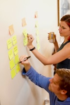 Two people are using sticky notes on a wall for organizing tasks or ideas. One person is writing on a note while the other observes with interest, holding a beverage can. The scene suggests collaborative planning or brainstorming.