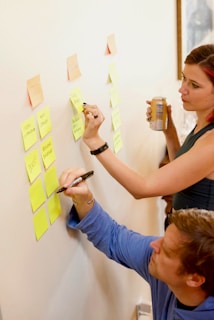Two people are using sticky notes on a wall for organizing tasks or ideas. One person is writing on a note while the other observes with interest, holding a beverage can. The scene suggests collaborative planning or brainstorming.
