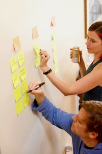 Two people are using sticky notes on a wall for organizing tasks or ideas. One person is writing on a note while the other observes with interest, holding a beverage can. The scene suggests collaborative planning or brainstorming.