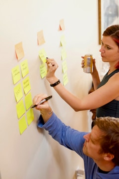 Two people are using sticky notes on a wall for organizing tasks or ideas. One person is writing on a note while the other observes with interest, holding a beverage can. The scene suggests collaborative planning or brainstorming.