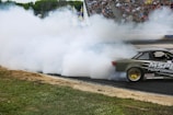 A sports car is drifting on a racetrack, producing a large cloud of white smoke. The vehicle is dark-colored with visible sponsorship decals, including 'MS Performance.' Spectators fill the stands in the background, watching the action. The track surface is visible near the car, and there is a grassy area adjacent to the track.