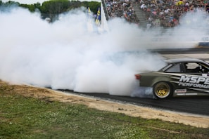 A sports car is drifting on a racetrack, producing a large cloud of white smoke. The vehicle is dark-colored with visible sponsorship decals, including 'MS Performance.' Spectators fill the stands in the background, watching the action. The track surface is visible near the car, and there is a grassy area adjacent to the track.