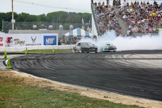 Two cars perform a drift around a racetrack surrounded by an audience of spectators in the stands. Smoke billows from the tires as the vehicles take a sharp turn. The track is marked with traffic cones, and the scene is lively with advertising banners along the walls.
