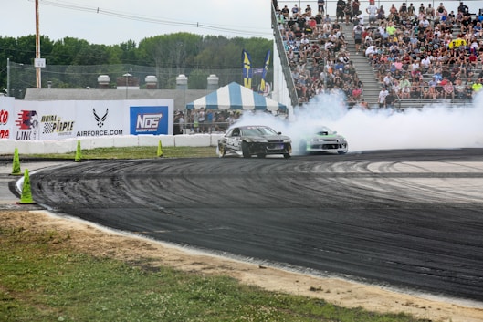 Two cars perform a drift around a racetrack surrounded by an audience of spectators in the stands. Smoke billows from the tires as the vehicles take a sharp turn. The track is marked with traffic cones, and the scene is lively with advertising banners along the walls.