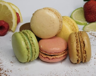 An arrangement of colorful macarons stacked together, featuring a variety of flavors. The image includes fresh fruit slices, such as lemon, lime, raspberry, and strawberry, enhancing the vibrant presentation. The background is a clean, white surface with a sprinkling of cocoa powder.