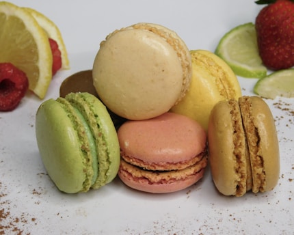 An arrangement of colorful macarons stacked together, featuring a variety of flavors. The image includes fresh fruit slices, such as lemon, lime, raspberry, and strawberry, enhancing the vibrant presentation. The background is a clean, white surface with a sprinkling of cocoa powder.