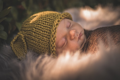 A sleeping baby is snugly wrapped in a cozy brown blanket and wearing a knitted green hat with ear-like extensions. The baby rests on a soft, furry surface, surrounded by a gentle, warm, natural light that enhances the peaceful atmosphere. The background features soft greenery, adding to the serene environment.