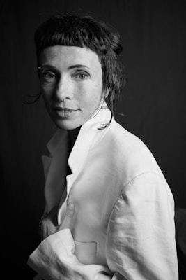A black-and-white portrait of a woman with short, dark hair and bangs. She is wearing a light-colored jacket and looking at the camera with a thoughtful expression. The lighting highlights the texture of her jacket and casts soft shadows on her face.
