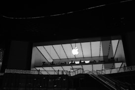 A large store with a glass facade is illuminated at night. The prominent logo of a well-known technology company is visible in the center. Inside, silhouettes of people can be seen near the windows, suggesting activity within. Exterior escalators and decorative lights are present in the foreground.