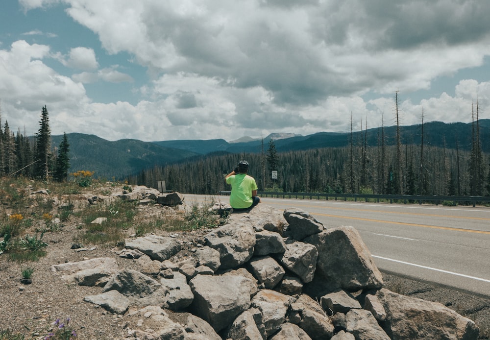 person in green hoodie sitting on gray rock near green trees during daytime