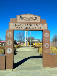 A war memorial with a red granite archway inscribed with 'Texas Panhandle War Memorial.' The structure features emblems and inscriptions related to various branches of the military. Behind the arch, a pathway leads to flagpoles, and small shrubs and trees line the memorial area under a clear blue sky.