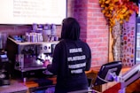 A person wearing a black hoodie with hashtags like #ORGANIC, #PLANTBASED, #SMOOTHIEBOWL, and #YUMMY stands at an espresso machine making a coffee. The background includes a menu, a wall with an autumn-themed decoration, and various equipment like a cash register.