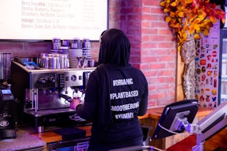 A person wearing a black hoodie with hashtags like #ORGANIC, #PLANTBASED, #SMOOTHIEBOWL, and #YUMMY stands at an espresso machine making a coffee. The background includes a menu, a wall with an autumn-themed decoration, and various equipment like a cash register.