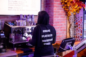 A person wearing a black hoodie with hashtags like #ORGANIC, #PLANTBASED, #SMOOTHIEBOWL, and #YUMMY stands at an espresso machine making a coffee. The background includes a menu, a wall with an autumn-themed decoration, and various equipment like a cash register.