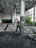 A person is standing in an abandoned industrial building. The space is dilapidated, with peeling paint on the walls and exposed structural elements. There is debris scattered across the floor and signs of neglect, such as overgrown plants visible through the broken windows. The atmosphere is one of decay and abandonment.