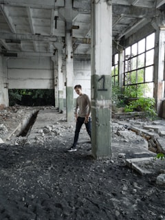 A person is standing in an abandoned industrial building. The space is dilapidated, with peeling paint on the walls and exposed structural elements. There is debris scattered across the floor and signs of neglect, such as overgrown plants visible through the broken windows. The atmosphere is one of decay and abandonment.