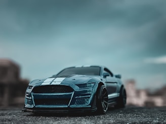 A model car sits prominently on a rough surface, featuring a sleek design with two white racing stripes running along the hood. The blurred background creates a sense of depth and the overall image is captured in low light, giving it a moody and dramatic atmosphere.