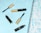 Several open bottles of mascara are scattered on a light blue geometric background with a subtle water pattern. The mascara tubes are black with gold accents, elegantly showcasing their sleek, modern design.