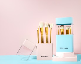 A set of golden-handled makeup brushes displayed in stylish white and blue holders with the brand name 'MISS SWISS'. The brushes have soft, white bristles and are neatly arranged in the containers on a pastel-colored background.