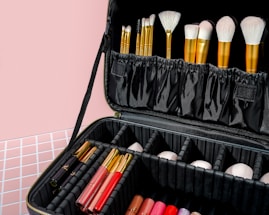 A black makeup case filled with various makeup brushes in gold handles is prominently displayed. The case also contains multiple compartments housing lipsticks and other makeup products arranged neatly. The background features a pastel pink wall and a patterned surface.