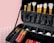 A black makeup case filled with various makeup brushes in gold handles is prominently displayed. The case also contains multiple compartments housing lipsticks and other makeup products arranged neatly. The background features a pastel pink wall and a patterned surface.