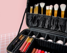 A black makeup case filled with various makeup brushes in gold handles is prominently displayed. The case also contains multiple compartments housing lipsticks and other makeup products arranged neatly. The background features a pastel pink wall and a patterned surface.