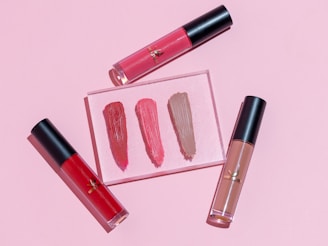 Three tubes of liquid lipstick are arranged beside swatches on a pink background. The lipsticks have transparent containers with black caps, showcasing shades of red, pink, and nude.