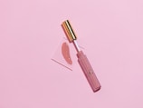 A makeup lip gloss with a gold cap lies on a soft pink background. The gloss is slightly opened and the applicator is placed on a small transparent square with a swatch of the lip gloss color beside it.